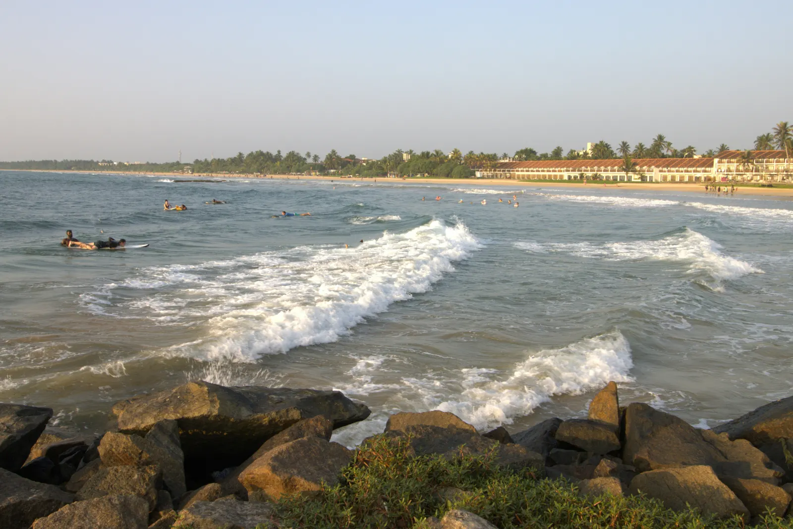 Waves rolling in over rocks toward the long curve of Bentota beach, with swimmers and palms in the distance